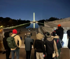 A group of people stand facing the illuminated Washington Monument at night, with its reflection visible in the Reflecting Pool.