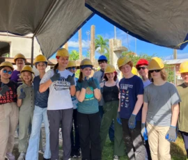 A group of teenagers wearing yellow hard hats and work gloves gather under a tarp at a construction site, smiling at the camera on a sunny day.