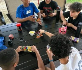 Five teenagers sit around a table outdoors, playing a game of UNO. Cards are spread on the table, and various drinks are nearby.