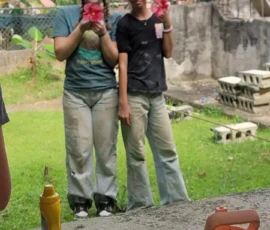 Two people stand outside holding drinks, with a table in the foreground displaying plates with food, water bottles, and a bottle of mustard. Green grass and banana trees are in the background.