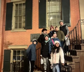 A group of children in winter clothing stand on the steps outside a brick building with green shutters at night.