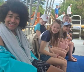 A group of young people sit on a boat, some smiling at the camera, with trees and more people visible in the background.