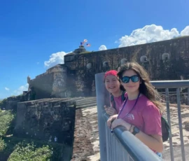Two people in pink shirts stand by a railing in front of an old stone fort with a flag, under a blue sky with scattered clouds.