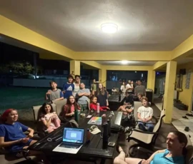 A group of people sit and stand around tables in a brightly lit covered outdoor area at night, with laptops and drinks visible on the tables.
