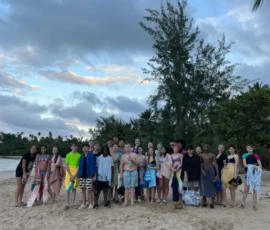 A group of teenagers in swimsuits and towels stand together on a sandy beach with trees in the background under a cloudy sky.