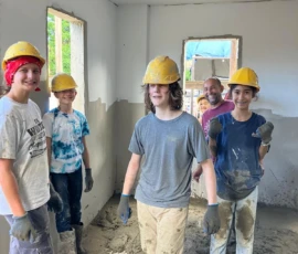 Five people wearing yellow hard hats and work gloves stand inside a partially constructed building with unfinished walls and windows, smiling at the camera.