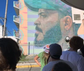 A group of people stand in front of a large mural painted on a building, depicting a man's face in profile with a beard, earring, and cap.