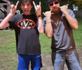 Two people stand outside at night on grass, both making hand gestures and posing playfully for the camera. One wears a black T-shirt and shorts, the other sunglasses, a hat, and jeans.