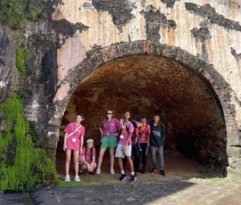 A group of six people stand and crouch under a large stone archway with moss and weathered brick walls at an old fort.