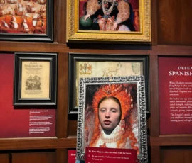 A child poses with their face in a Queen Elizabeth I cutout, holding a red speech board, beneath a framed portrait of the queen in a museum exhibit.