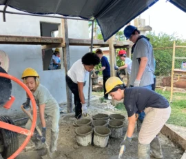 A group of people wearing hard hats and boots mix and shovel wet cement into buckets at a construction site under a tarp.