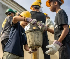 A group of people wearing yellow safety helmets and gloves work together, passing a bucket while standing on a construction site.