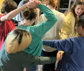 A group of six people stand in a circle indoors, holding hands and interlocking arms in a tangled manner as if playing a team-building game.