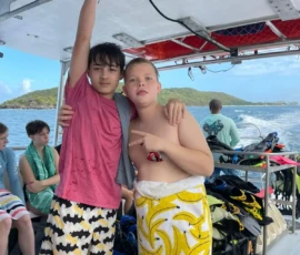 Two boys in swim trunks stand together on a boat, one with his arm raised and the other pointing, with other kids seated in the background and ocean scenery visible.