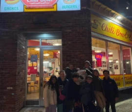 A group of people poses for a photo at night outside Ben's Chili Bowl restaurant, which is brightly lit with neon signs.