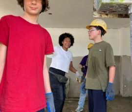 A group of people wearing hard hats, gloves, and casual clothes stand indoors at a construction or renovation site.