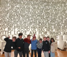 A group of eight children pose in front of a large wall covered with white three-dimensional words and phrases arranged in rows.
