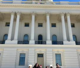 A group of people are sitting on the steps in front of the U.S. Capitol building under a clear blue sky.