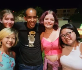 Seven people stand close together outdoors at night, smiling and posing for a group photo. Buildings and greenery are visible in the background.