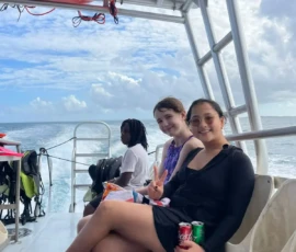 Three people sit on a boat; two smile at the camera, one holds a can of soda and makes a peace sign. Scuba gear and the ocean are visible in the background.
