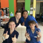 Four people in blue scrubs pose indoors, two giving thumbs up and two making peace signs, with colorful walls and tables in the background.