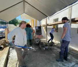Four people work under a canopy at a construction site, mixing materials and filling buckets, with tools and equipment scattered around.