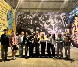 A group of eleven people stands side by side in front of a colorful mural featuring civil rights imagery and prominent figures inside an art gallery.