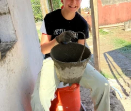 A person wearing a yellow hard hat and gloves sits on an overturned bucket, holding a bucket filled with wet cement, outdoors next to a building.