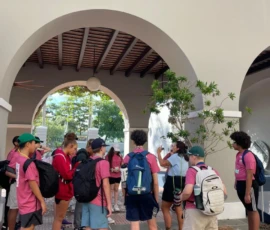 A group of students wearing pink shirts and backpacks stand under an archway, listening to a person speaking to them.