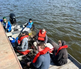 Five people wearing life jackets sit in a metal boat next to a wooden dock on a sunny day by a large body of water.