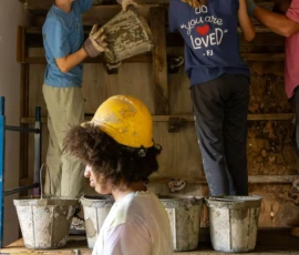 Several people wearing hard hats work with buckets of cement or plaster on scaffolding indoors; one person stands in the foreground.