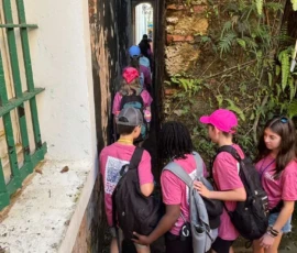 A group of children wearing pink shirts and backpacks walk in single file through a narrow alley lined by walls and plants.