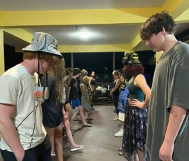 Two lines of people stand facing each other under a covered patio at night, some wearing casual clothes and hats, appearing to participate in a group activity.