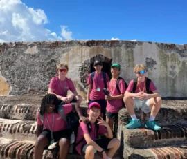 Six children in matching pink shirts sit and pose on stone steps in front of an old, weathered wall under a bright blue sky with some clouds.