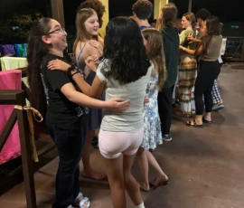 A group of teenagers stand in pairs facing each other, holding hands and talking on a covered patio at night.