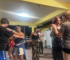 Group of people dancing in pairs indoors under yellow beams, with smiling faces and casual clothing.