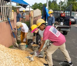 Four people wearing hard hats and gloves shovel gravel into buckets on a street next to a truck; a man stands nearby by a fence.
