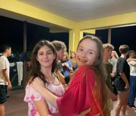 Two girls smile and pose together while dancing at a social event indoors, surrounded by other teenagers.