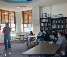 A woman stands at the front of a classroom holding a sign, while four students sit at tables listening. Bookshelves are visible in the background.