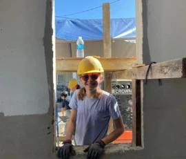 A person wearing a yellow hard hat, safety glasses, gloves, and a white shirt stands outside a window frame at a construction site on a sunny day.