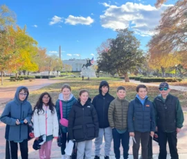 A group of eight children in winter jackets stand in a row outdoors with trees, the Washington Monument, and a statue visible in the background on a sunny day.