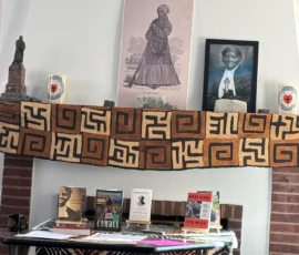 A mantle with framed portraits and statues of Harriet Tubman, patterned cloth, and a table displaying books and memorabilia dedicated to her.