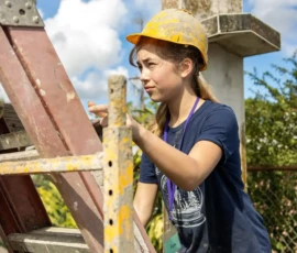 A woman wearing a yellow hard hat climbs a construction ladder outdoors on a sunny day.