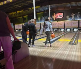 Three people stand at the edge of bowling lanes holding orange balls, while another person waits nearby at a bowling alley with a "Rock 'n Bowl" sign overhead.