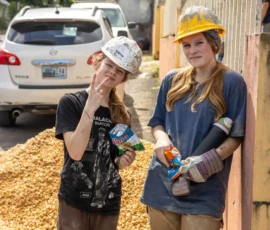Two young people wearing construction helmets and work clothes stand by a wall, holding snacks, with a parked car and a pile of gravel in the background.
