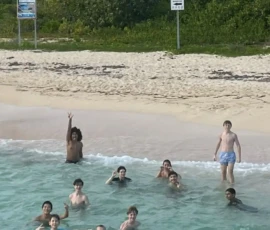 A group of children and teens swim in shallow ocean water near a sandy beach with greenery and signs in the background.