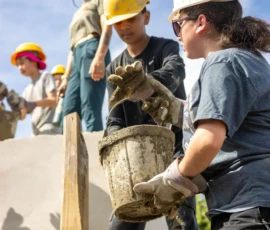 Several people wearing safety helmets and gloves work together on a construction site, passing a muddy bucket as they build a structure outdoors.