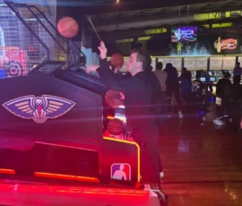 A person plays an arcade basketball game, aiming a ball at the hoop in a dimly lit game center with neon signs and other arcade machines in the background.