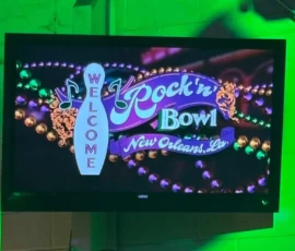 A television screen displays a welcome sign for Rock 'n' Bowl in New Orleans, Louisiana, with musical notes and colorful beads as decoration.