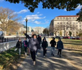 A group of people walk along a city sidewalk on a sunny day, with one person energetically jumping and reaching upward. Trees and buildings line the background.